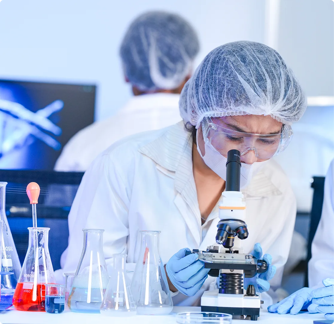 Femme scientifique regardant dans un microsope, avec des fioles, des béchers, des échantillons, et à l'arrière-plan, une autre personne qui travaille sur un ordinateur.